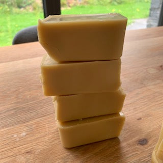 Four stacked rectangular bars of pale yellow wood polish on a wooden table, with a blurred window and green outdoor view in the background.