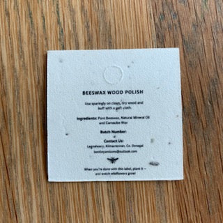 A square white paper label with black text reading 'BEESWAX WOOD POLISH' and product details, placed on a brown wood surface.