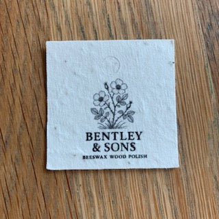 A square white paper label for Bentley & Sons Besswax Wood Polish, featuring a black floral illustration and text, placed on a wooden surface.