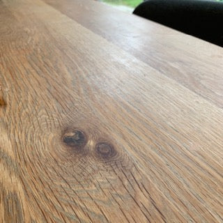 Close-up of a natural wood tabletop with visible grain, knots, and a warm medium-brown finish. Part of a dark chair is visible in the upper right corner.